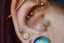 a boldly curated ear with stakce dlobe piercings, an industrial one, a tropical conch, a helix and a tragus piercing done with rhinestone gold studs, a bold blue one