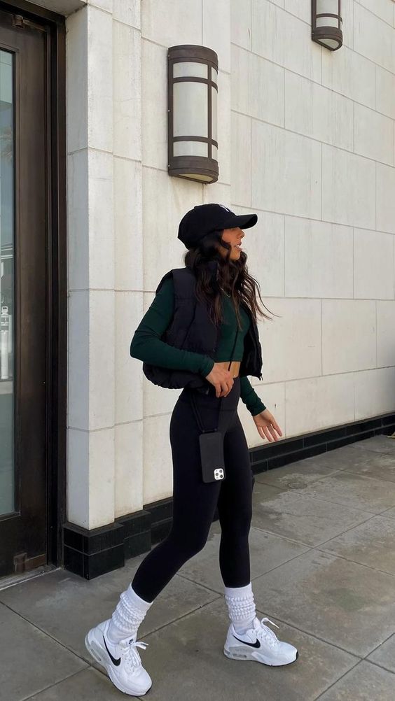 a sport chic outfit for spring - black leggings, a green long sleeve crop top, a black quilted vest, a black cap, white trainers