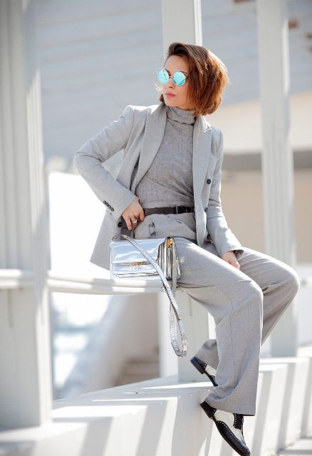With gray fitted turtleneck, light gray long blazer, light gray pants, black and white leather lace up flat shoes, black leather belt and mirrored rounded sunglasses