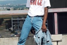 a 90s outfit with a white printed t-shirt, blue jeans, white socks and sneakers, a blue ripped denim jacket and a burgundy beanie