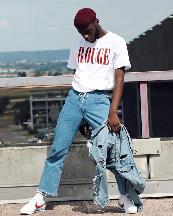 a 90s outfit with a white printed t-shirt, blue jeans, white socks and sneakers, a blue ripped denim jacket and a burgundy beanie