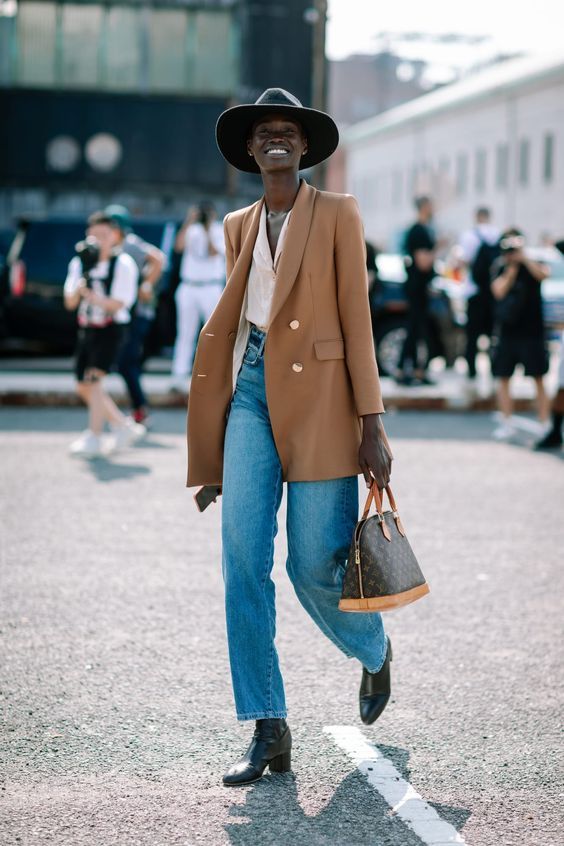 a bold and cool look with a white shirt, blue jeans, black boots, a camel oversized blazer, a printed bag and a black wide-brim hat