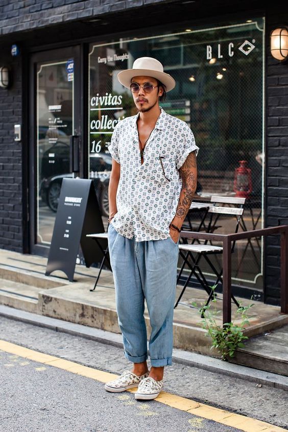 a summer boho look with a printed short-sleeved shirt, blue linen pants, printed snakers and a neutral hat