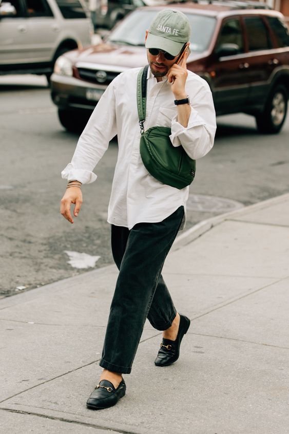 a white button down, black jeans, black loafers, a green waist bag and a green baseball cap