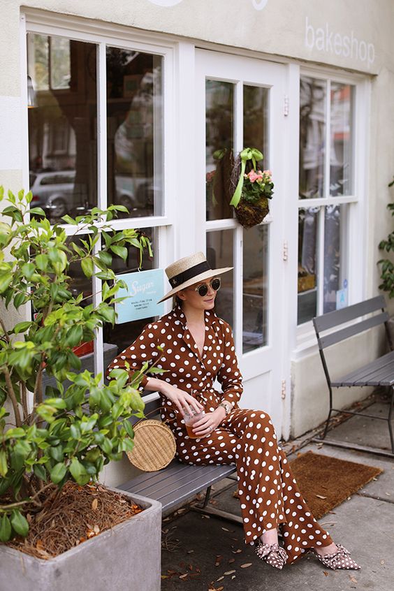 a refined and chic vacation look with a rust colored jumpsuit and matching shoes with bows, a straw hat and a woven bag