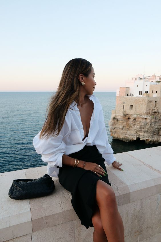 a refined vacation look with an oversized white button down, a black slip midi skirt, statement accessories and a black clutch