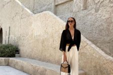 a vacation outfit with a neutrla bre and linen pants, a black knotted shirt, black slides and a woven bag