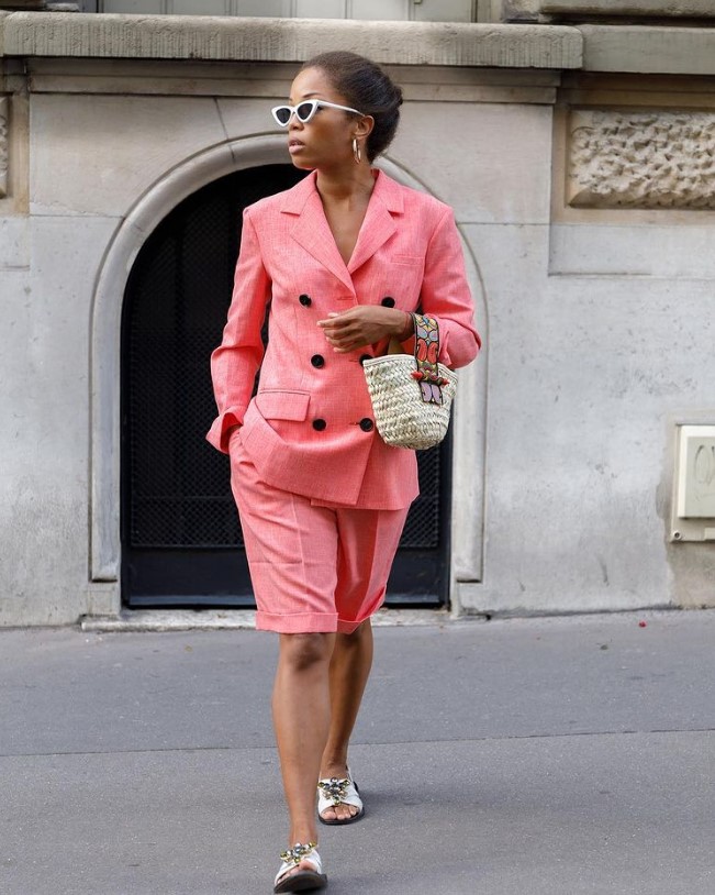 a coral pink linen suit with a blazer and Bermuda shorts, a woven bag, white slides and white frame sunglasses