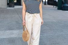 With beige and black wide brim hat, rounded sunglasses, black and white striped loose shirt, beige straw rounded bag and beige sandals