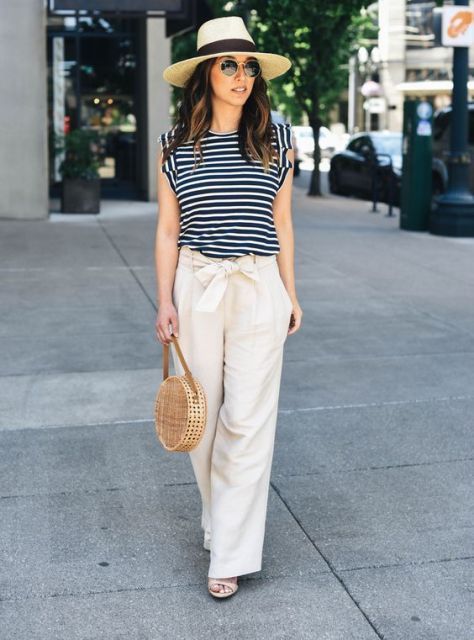 With beige and black wide brim hat, rounded sunglasses, black and white striped loose shirt, beige straw rounded bag and beige sandals