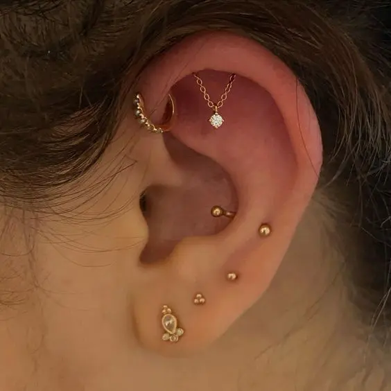 a bold ear look with a forward helix, a hidden helix, sung, lobe piercings done with gold and rose gold studs and a unique chain piece