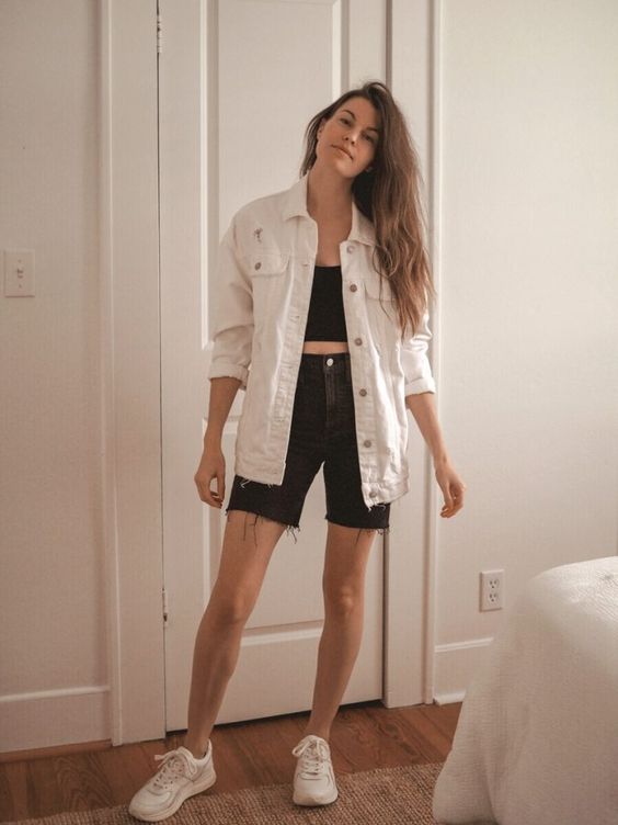 a casual summer outfit with a black crop top, black denim shorts, a white oversized denim jacket and white trainers
