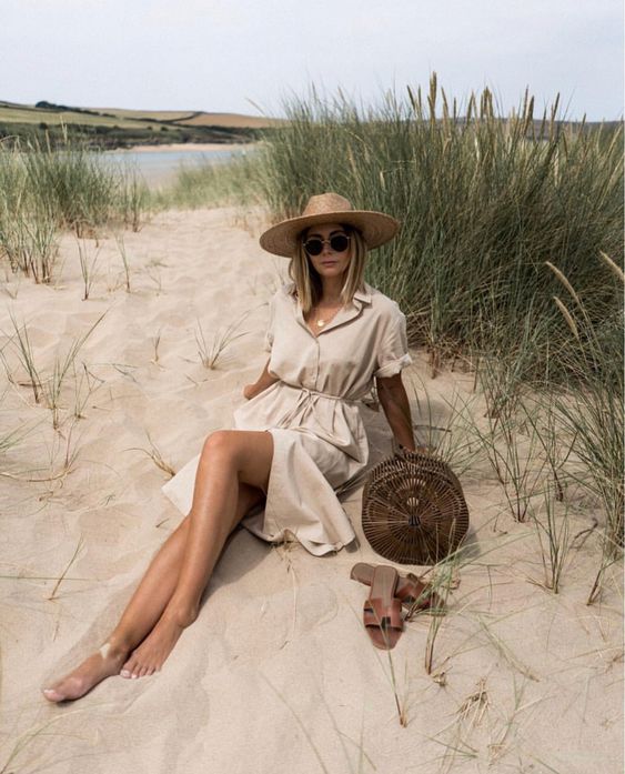 a neutral linen midi dress with buttons, a wide brim straw hat, a rattan round bag and brown slides