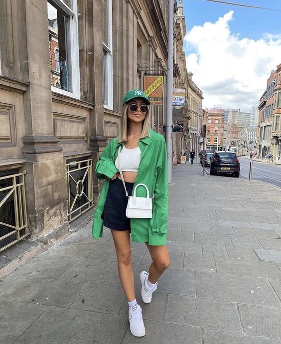a sport chic outfit with a white crop top, black shorts, a bold grene oversized shirt, white trainers and socks, a white bag and a green baseball cap