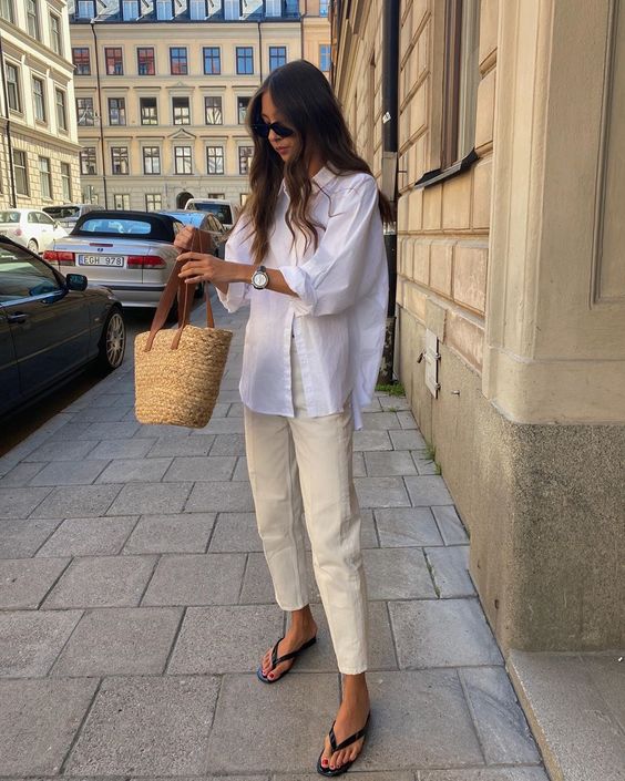 a neutral summer outfit with an oversized white shirt, neutral jeans, flipflops and a woven bag