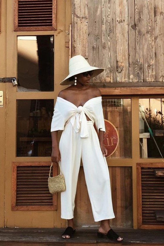 a stylish vacation look with a white off the shoulder knotted crop top and matching pants, black slippers and a hat