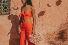 an orange linen suit with a knotted crop tpo and pants, black sandals and a colorful bag of wood