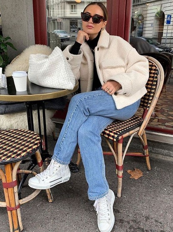 a black turtleneck, a creamy faux fur jacket, blue jeans, white sneakers and a creamy woven clutch is cool