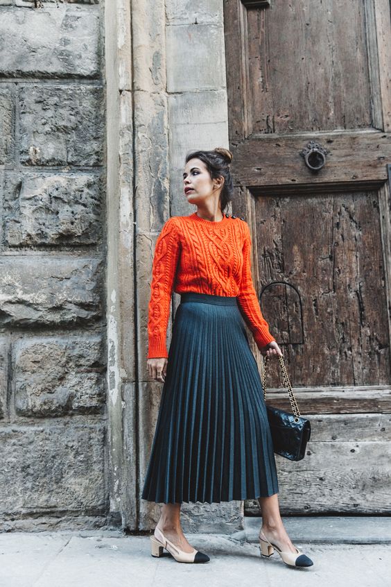 a bold fall outfit with an orange patterned sweater, a navy pleated midi, two-tone shoes and a black bag