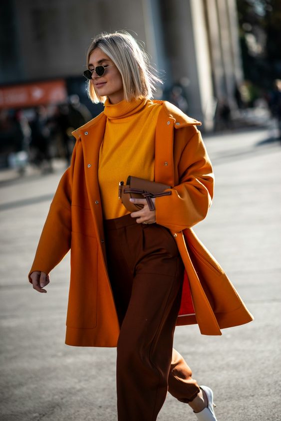 a fall tone work outfit with a yellow turtleneck, brown trousers, an orange coat, a brown bag and white shoes