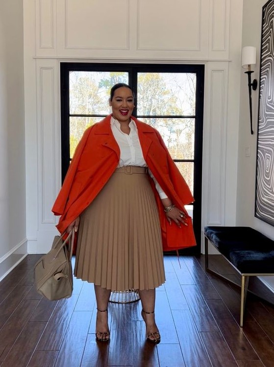 a neutral fall work outfit with a white shirt, a beige pleated midi, nude shoes and a bag accented with a bold orange coat