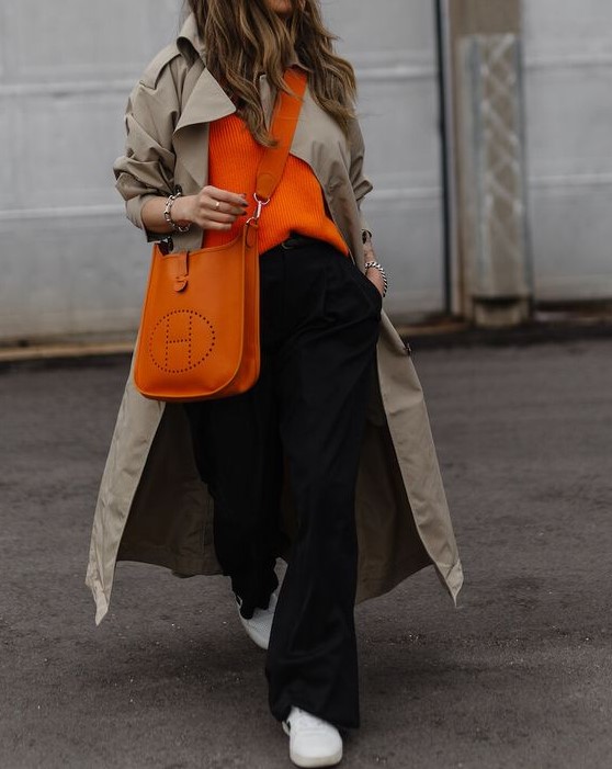 a comfortable casual look with an orange jumper, black trousers, a neutral trench, an orange tote and white sneakers