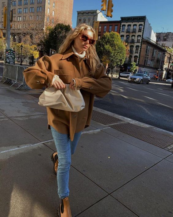a white sweater, a brown shacket, blue jeans, beige Chelsea boots and a creamy clutch for Thanksgiving