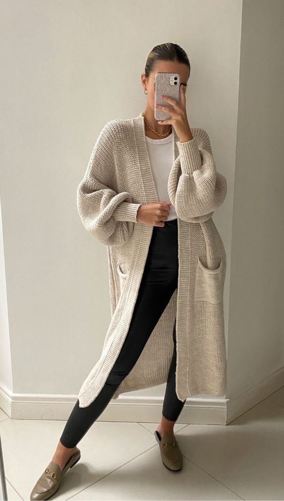 a simple and cozy Thanksgiving outfit with a white top, black leggings, a creamy midi cardigan, grey slippers and a necklace