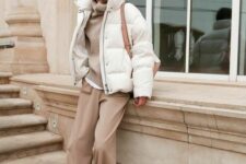 a minimalist winter outfit with a white t-shirt and a tan sweater over it, beige pants, white sneakers, a white puffer jacket and a beige bag