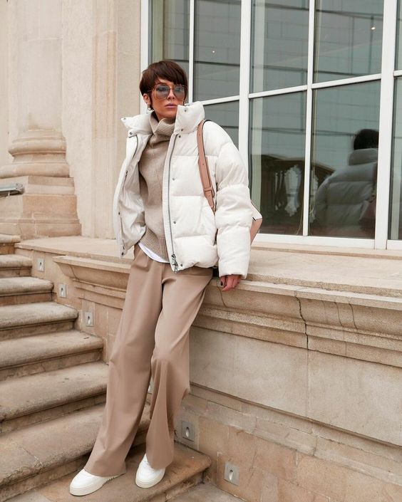 a minimalist winter outfit with a white t shirt and a tan sweater over it, beige pants, white sneakers, a white puffer jacket and a beige bag
