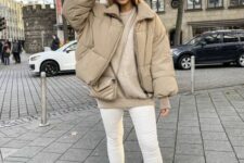 a neutral winter outfit with a greige sweater, a beige cropped puffer jacket, white jeans and boots, a coral beanie