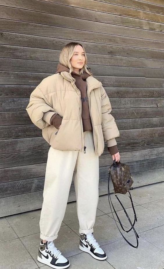 a sport chic outfit with a brown hoodie, neutral jeans, black and white trainers, a tan puffer jacket and a brown backpack