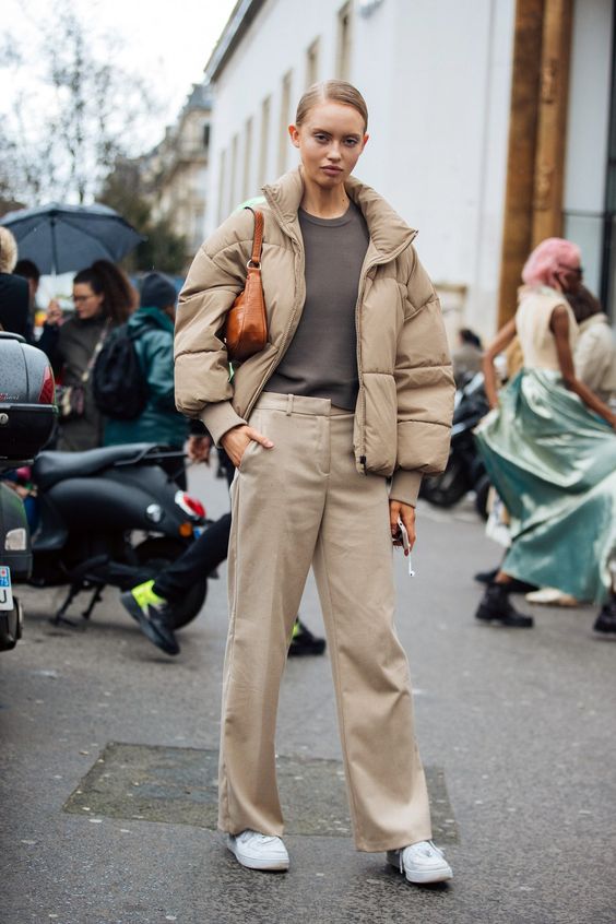 a stylish winter outfit with a taupe t shirt, beige trousers, a beige puffer jacket, white sneakers and an amber bag