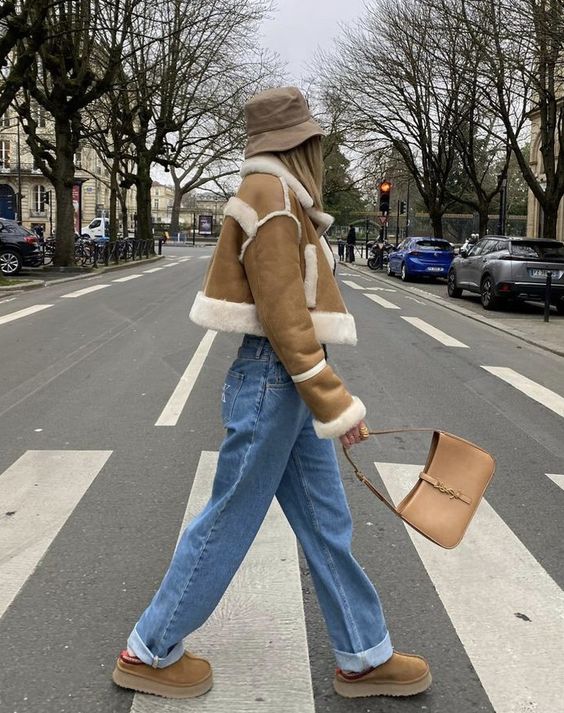 a cute and cozy winter outfit with high waisted blue jeans, camel ugg boots and a cropped aviator, a beige bag and a bucket hat