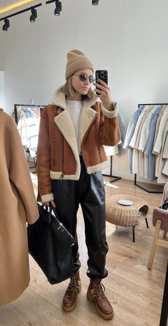 a sporty winter outfit with a white t-shirt, black leather pants, brown boots, a brown aviator, a beige beanie and a black bag