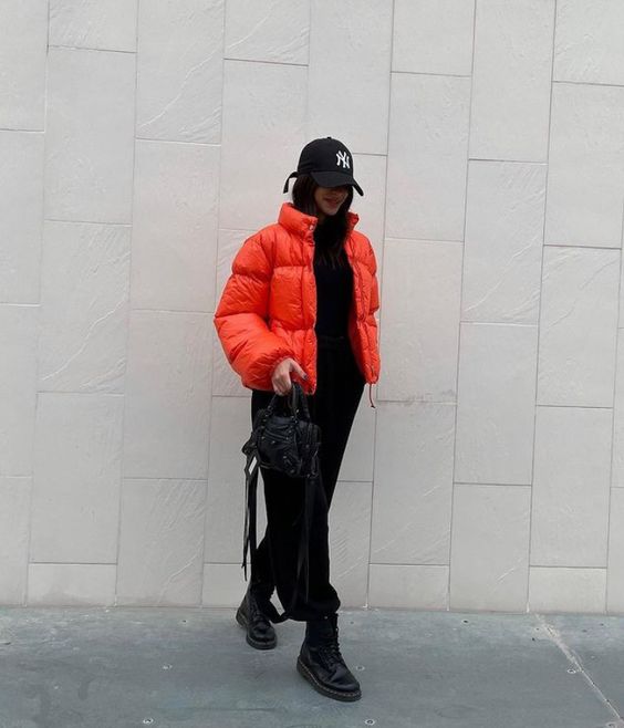 a total black outfit with a sweater and sweatpants, combat boots, an orange puffer and a black bag plus a baseball cap