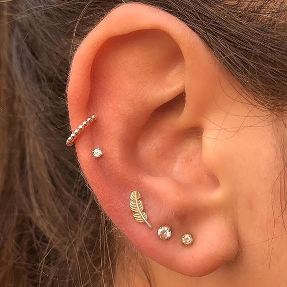 a triple lobe piercing done with rhinestone studs and a feather, a double mid helix piercing with a stud and a hoop