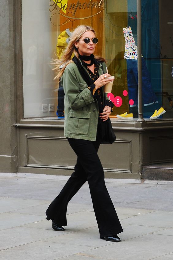 Kate Moss wearing a black floral blouse, a black scarf and flare pants, a green utility jacket and a black tote
