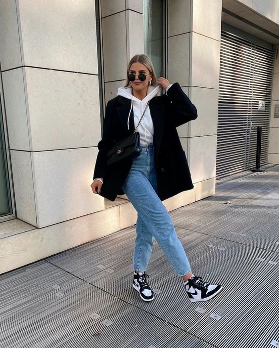a white hoodie, blue high waisted jeans, black and white trainers, a black blazer and a black bag for a sporty chic look