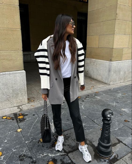 a white t shirt, black jeans, a grey oversized blazer, neutral trainers, a black bag and a Breton stripe sweater over the shoulders