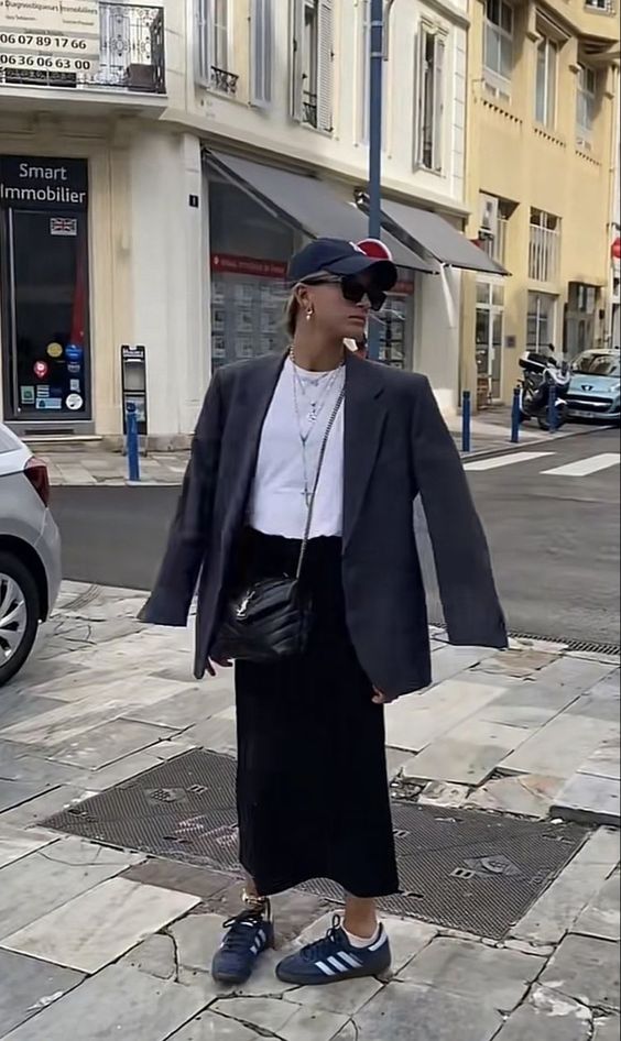 a comfy modern outfit with a black maxi slip skirt, a white t-shirt, an oversized grey blazer, navy Gazelle sneakers, a black bag and a cap