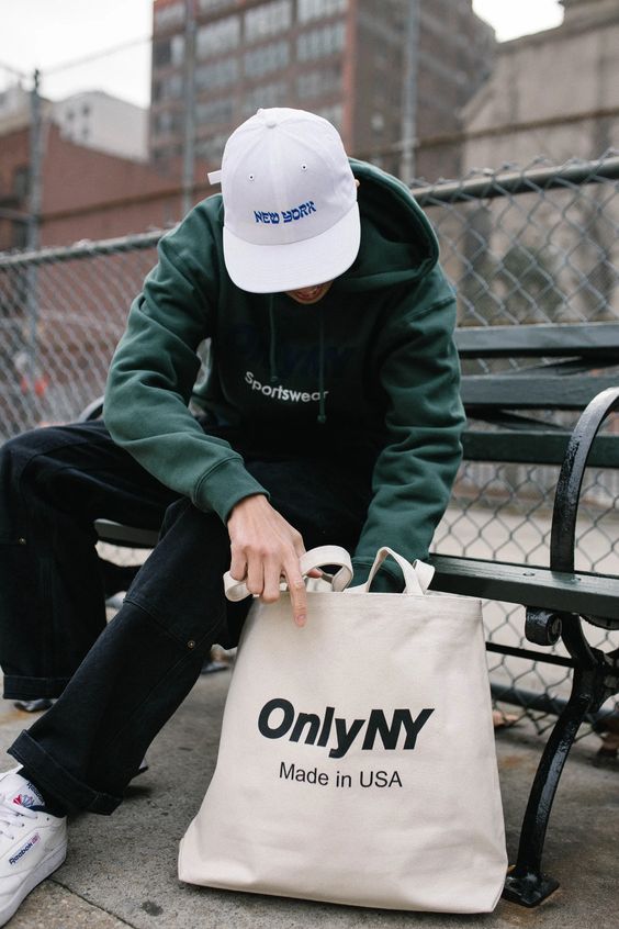 a sport outfit with a green hoodie, black jeans, white sneakers and socks, a white cap and a neutral canvas bag