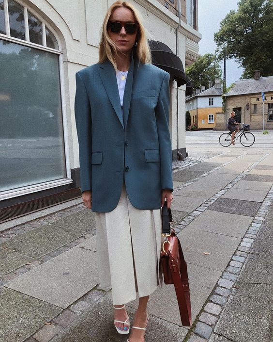 a white top, a neutral pleated midi skirt, white strappy heels, an oversized blue blazer and a brown bag for a chic spring to summer look