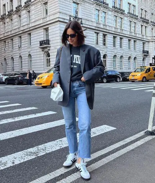 a black printed t-shirt, an oversized grey blazer, blue cropped jeans, white Samba sneakers and a bag on chain