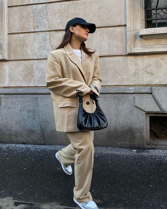 a white t shirt, a tan pantsuit with an oversized blazer, neutral trainers, a black cap and a black hobo bag
