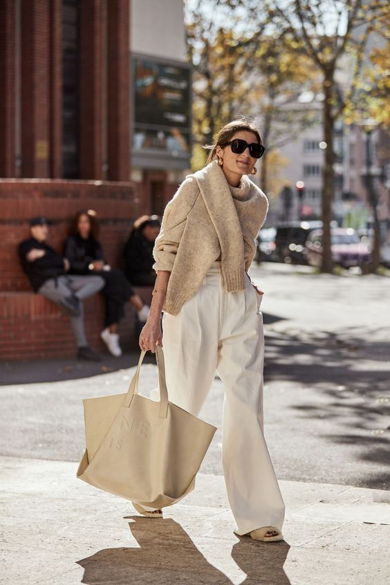 a neutral outfit with white high-waisted pants, neutral slippers, a tan sweater, a neutral canvas tote for a cold day