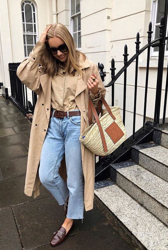 a beige shirt, a beige trench, blue jeans, a brown belt, brown loafers and a straw bag are a cool look for spring