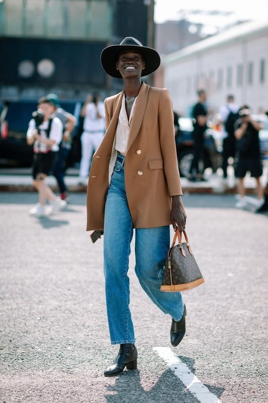 a bold and cool look with a white shirt, blue jeans, black boots, a camel oversized blazer, a printed bag and a black wide-brim hat