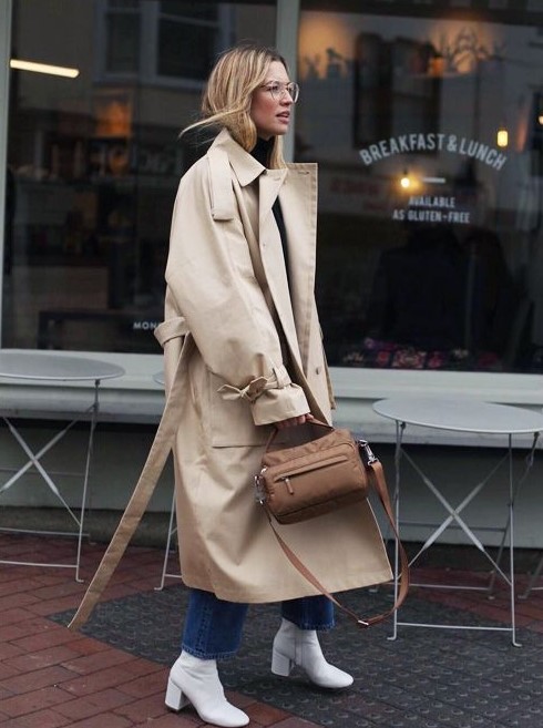 a stylish work look with a black turtleneck, blue jeans, white boots, a tan trench and a brown bag