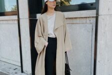 31 a tan t-shirt, black trousers, a beige maxi trench, grey trainers, a white cap and a black tote for an everyday look